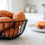 Whole raw sweet potatoes sit in a black wire basket on a bright white marble kitchen counter, with a plate of cooked, halved sweet potatoes softly blurred in the background.