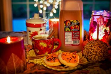 a festive fall display with a jug of apple cider, a mug of hot cider, and decorative treats and candles on a table top