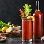 Bloody Mary cocktail in a condensation-covered glass with a spice-rimmed edge, garnished with celery, bacon, olives, pickles, and lemon, beside a tall, unlabeled glass bottle of red tomato-based Bloody Mary mix with a black cap, on a dark stone countertop with fresh celery, olives, lemon, and bacon arranged nearby against a charcoal background.