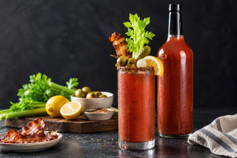 Bloody Mary cocktail in a condensation-covered glass with a spice-rimmed edge, garnished with celery, bacon, olives, pickles, and lemon, beside a tall, unlabeled glass bottle of red tomato-based Bloody Mary mix with a black cap, on a dark stone countertop with fresh celery, olives, lemon, and bacon arranged nearby against a charcoal background.