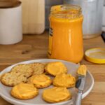 Open glass jar of orange Cheez Whiz cheese spread with its yellow lid beside a plate of golden crackers, several topped with the smooth cheese spread, and a butter knife on a wooden kitchen counter.