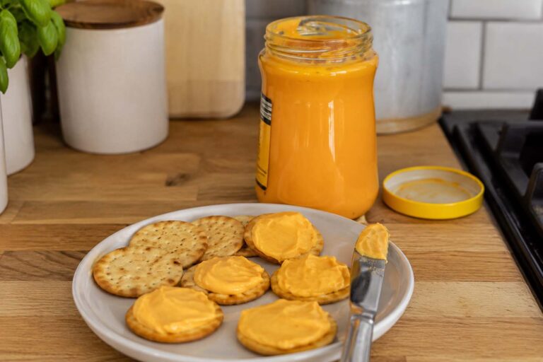 Open glass jar of orange Cheez Whiz cheese spread with its yellow lid beside a plate of golden crackers, several topped with the smooth cheese spread, and a butter knife on a wooden kitchen counter.