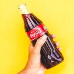 close up of a hand holding an open bottle of coca cola against a bright yellow background
