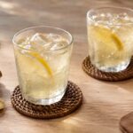 two fizzy glasses of ginger ale on a table with coasters, fresh ginger root, and lemon wedges