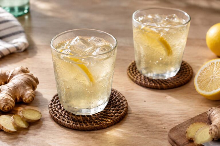 two fizzy glasses of ginger ale on a table with coasters, fresh ginger root, and lemon wedges