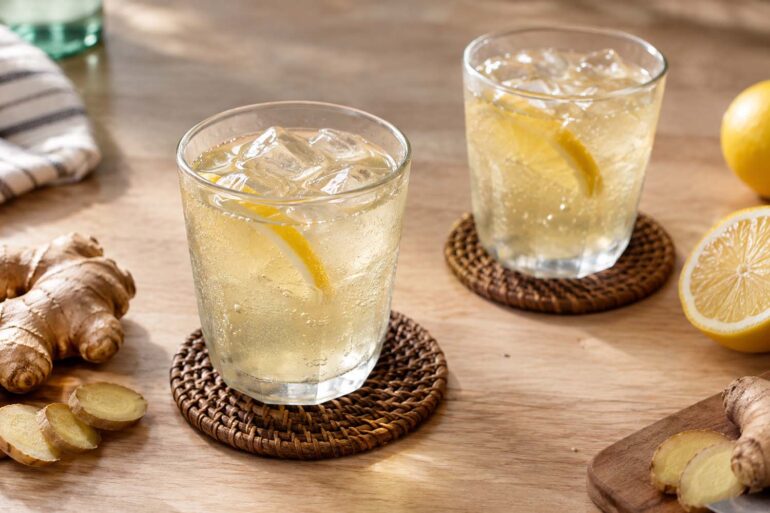two fizzy glasses of ginger ale on a table with coasters, fresh ginger root, and lemon wedges