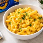 A white bowl of creamy, golden-orange Kraft macaroni and cheese topped with fresh green herbs and black pepper, with a Kraft macaroni and cheese box and an opened cheese powder packet on a light kitchen countertop nearby.