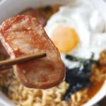 an up close shot of a slice of fried spam held with chopsticks above rice and a fried egg