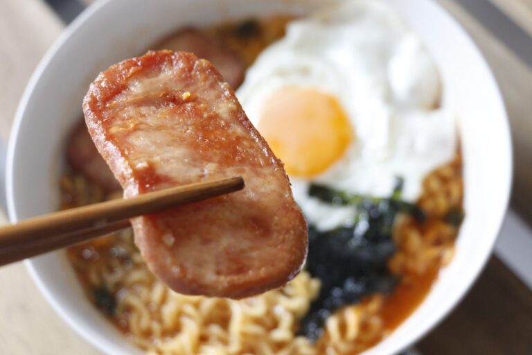 an up close shot of a slice of fried spam held with chopsticks above rice and a fried egg