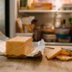 close-up of an opened rectangular block of Velveeta cheese resting on its silver foil wrapper on a wooden cutting board. A small kitchen knife and a few sliced pieces of cheese sit beside the main block