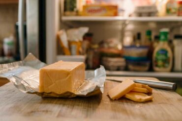 close-up of an opened rectangular block of Velveeta cheese resting on its silver foil wrapper on a wooden cutting board. A small kitchen knife and a few sliced pieces of cheese sit beside the main block