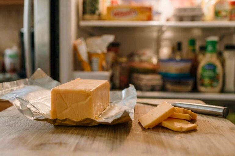 close-up of an opened rectangular block of Velveeta cheese resting on its silver foil wrapper on a wooden cutting board. A small kitchen knife and a few sliced pieces of cheese sit beside the main block