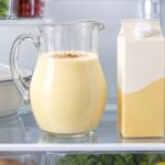 A glass pitcher of homemade eggnog sprinkled with nutmeg sits on a refrigerator shelf beside a plain store-bought eggnog carton and a covered white casserole dish in a clean, brightly lit refrigerator.