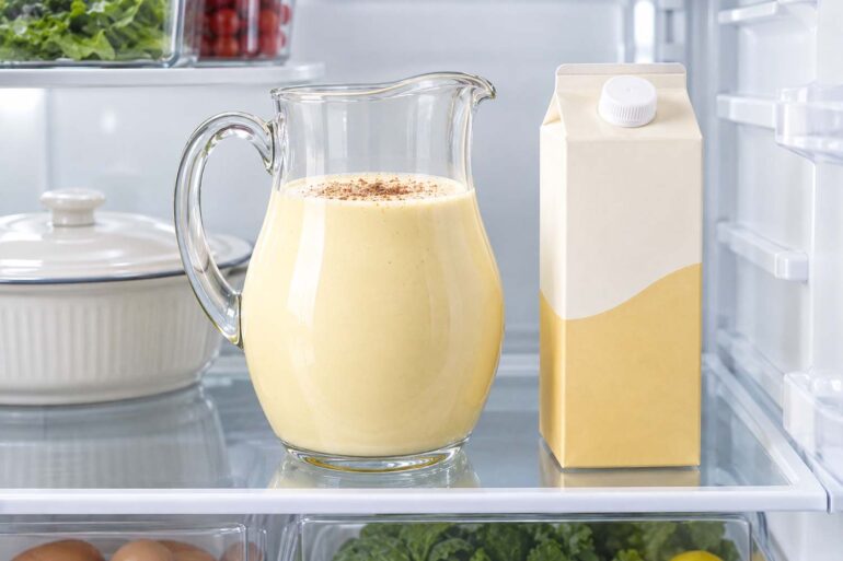 A glass pitcher of homemade eggnog sprinkled with nutmeg sits on a refrigerator shelf beside a plain store-bought eggnog carton and a covered white casserole dish in a clean, brightly lit refrigerator.
