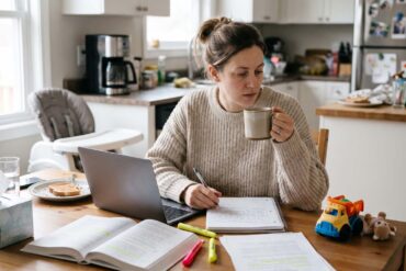 A 35-year-old mother wearing a cozy sweater studies on her laptop at the kitchen table in the early morning, with a notebook, coffee mug, and a child's toy nearby.