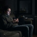 A moody photo of a man sitting alone on a couch in a dark room at night. His face is illuminated only by the soft glow of his smartphone screen as he scrolls, conveying a sense of quiet isolation.