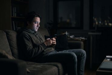 A moody photo of a man sitting alone on a couch in a dark room at night. His face is illuminated only by the soft glow of his smartphone screen as he scrolls, conveying a sense of quiet isolation.