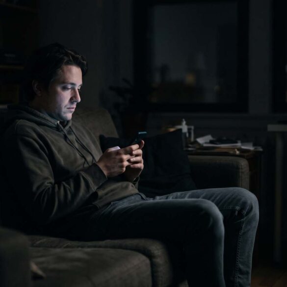 A moody photo of a man sitting alone on a couch in a dark room at night. His face is illuminated only by the soft glow of his smartphone screen as he scrolls, conveying a sense of quiet isolation.