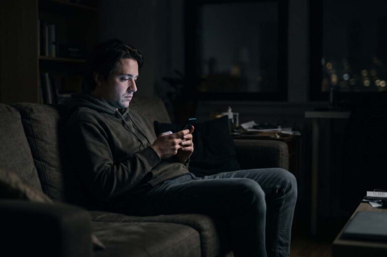 A moody photo of a man sitting alone on a couch in a dark room at night. His face is illuminated only by the soft glow of his smartphone screen as he scrolls, conveying a sense of quiet isolation.