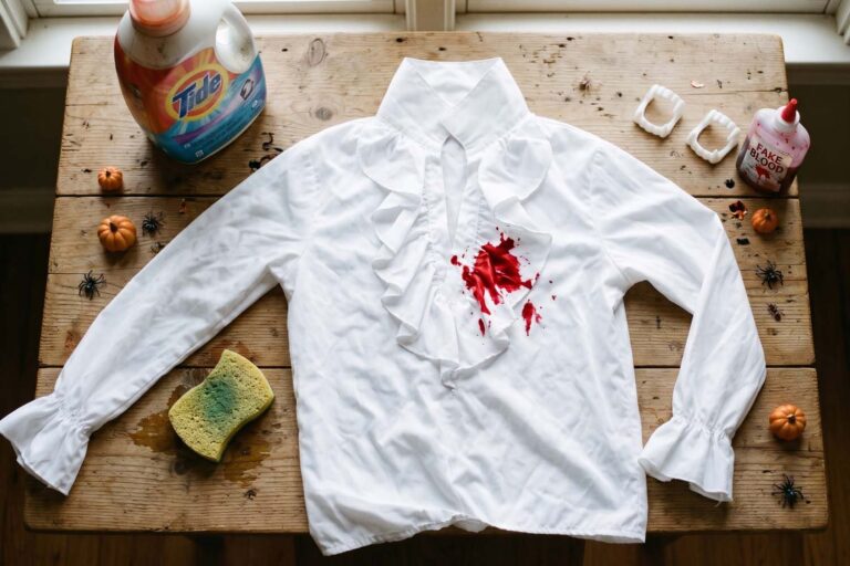 A white ruffled Halloween costume shirt with a bright red fake blood stain lies flat on a rustic wooden table. The shirt is surrounded by stain-removal supplies, including a Tide laundry detergent bottle and a damp sponge, alongside a bottle of fake blood, vampire fangs, and small Halloween decorations like spiders and mini pumpkins.