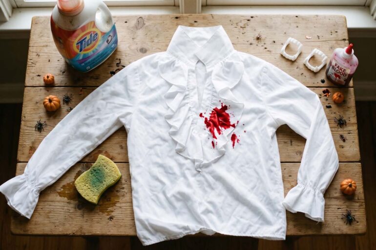 A white ruffled Halloween costume shirt with a bright red fake blood stain lies flat on a rustic wooden table. The shirt is surrounded by stain-removal supplies, including a Tide laundry detergent bottle and a damp sponge, alongside a bottle of fake blood, vampire fangs, and small Halloween decorations like spiders and mini pumpkins.