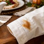 A brown gravy stain on the cuff of an ivory cotton shirt beside a gravy boat and holiday dinner table.