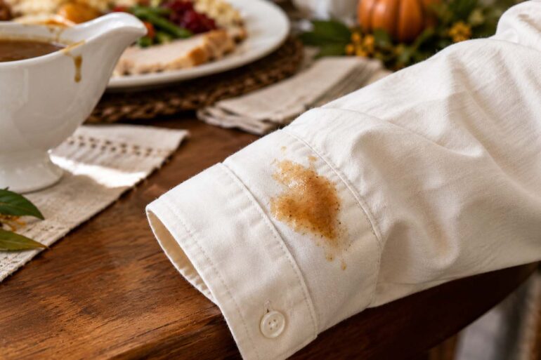 A brown gravy stain on the cuff of an ivory cotton shirt beside a gravy boat and holiday dinner table.