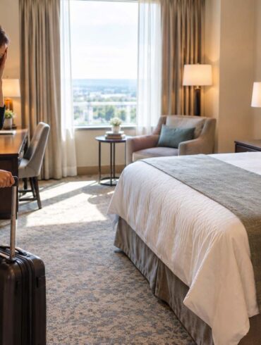 A weary traveler stands in the doorway of an upscale hotel room with one rolling suitcase, resting a hand against her forehead in frustration after arriving. The room features a neatly made bed, warm neutral furnishings, a sitting area, soft carpet, bedside tables, and natural daylight from a large window.