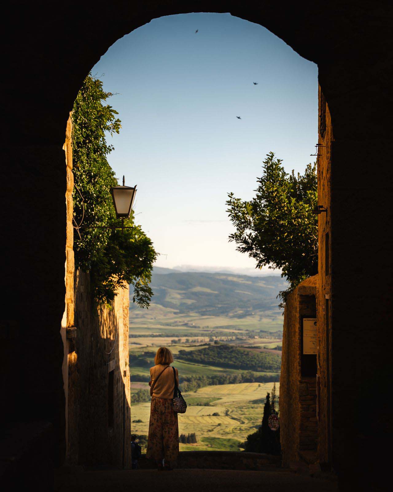 A woman stands under an arched passage, looking out onto a beautiful countryside landscape with clear skies.