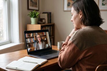 An adult sits at a wooden desk in a comfortably lit home, viewed from over their shoulder. They are attentively engaged in a virtual mental health appointment on their laptop. The screen displays a professional psychiatric provider on a video call in an office setting. An open notebook and a pen rest on the desk next to the computer, illuminated by soft natural light from a nearby window.