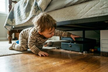 A toddler in pajamas crawling on a wooden floor, reaching under a bed toward a dark, partially open lockbox.