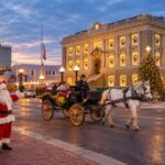 Santa waves beside a horse-drawn holiday carriage on a festive downtown street at dusk, with a historic clock-fronted building illuminated by Christmas lights, decorated windows, a towering Christmas tree, glowing streetlamps, and wet pavement reflecting the warm holiday lights.