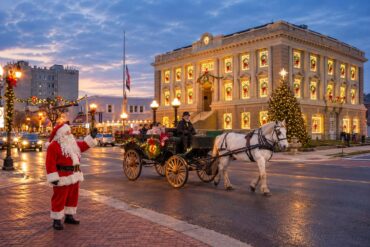 Santa waves beside a horse-drawn holiday carriage on a festive downtown street at dusk, with a historic clock-fronted building illuminated by Christmas lights, decorated windows, a towering Christmas tree, glowing streetlamps, and wet pavement reflecting the warm holiday lights.