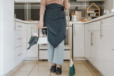 a woman standing in a kitchen holding a broom and cleaning supplies