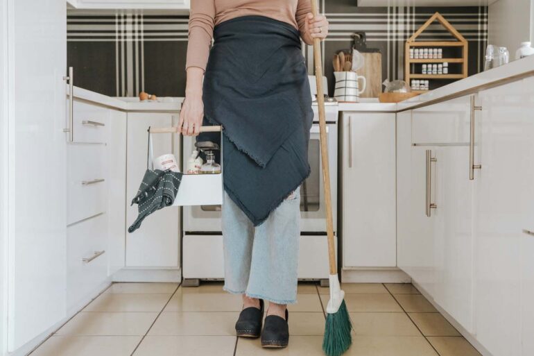 a woman standing in a kitchen holding a broom and cleaning supplies