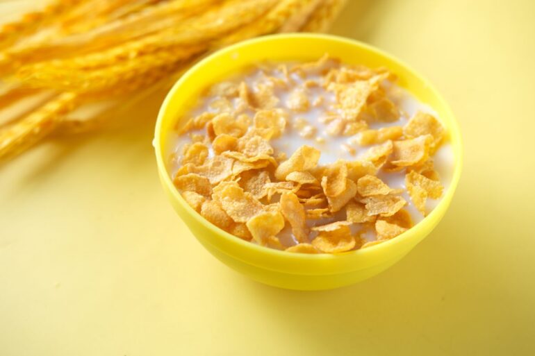 A bowl of corn flakes with milk on a table next to wheat husks